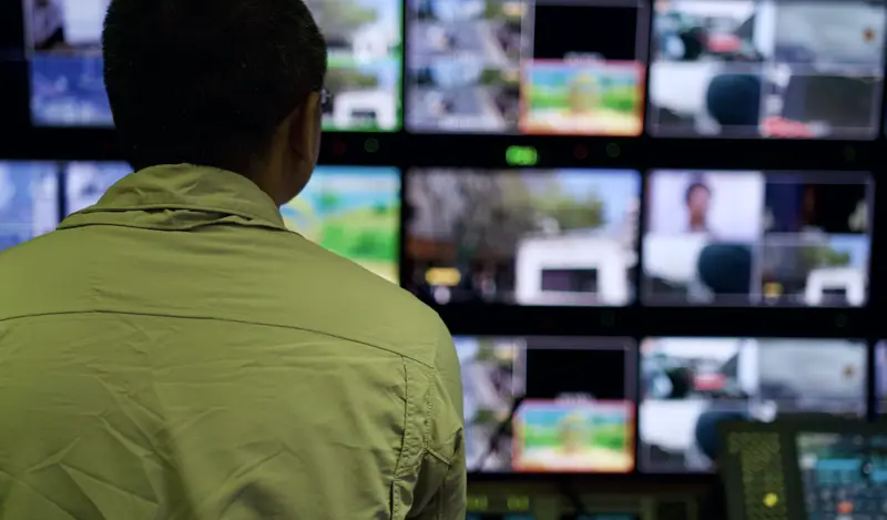 Operator facing a wall of broadcast monitoring screens in a dark control room